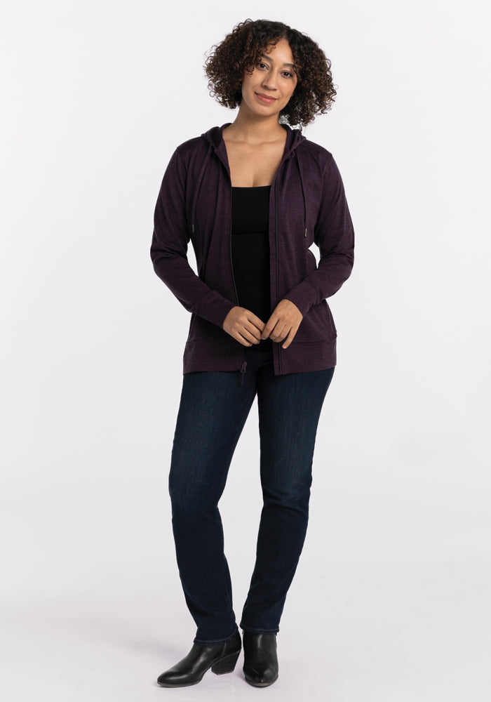 A person with curly hair is standing facing the camera, wearing the Woolx Ryann Hoodie in Deep Plum, which features a lightweight and athletic fit over a black top. They are complemented by dark jeans and black boots, with a plain white background accentuating their style.