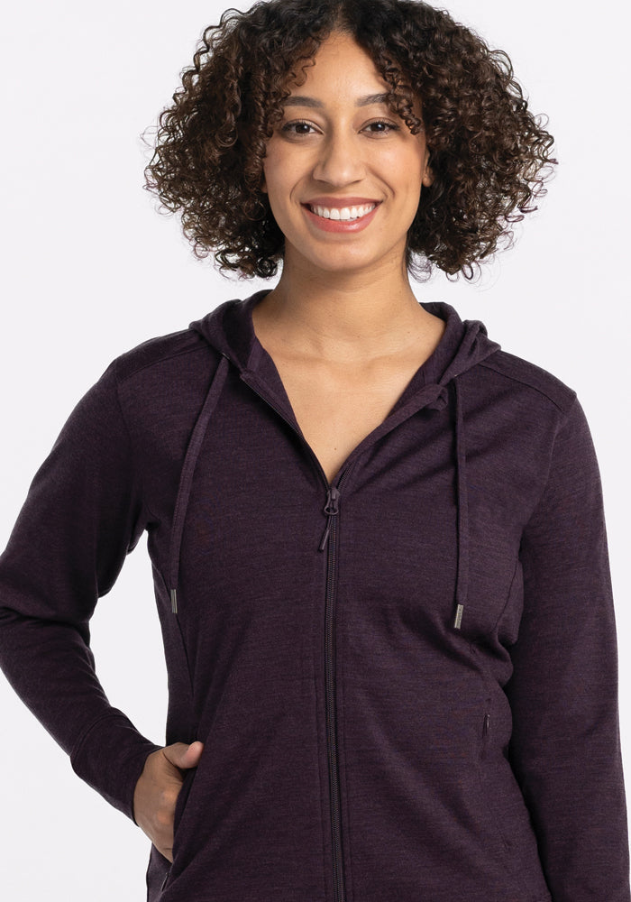 A person with curly hair smiles while wearing a Woolx Ryann Hoodie in deep plum, featuring an athletic fit. The background is plain white.