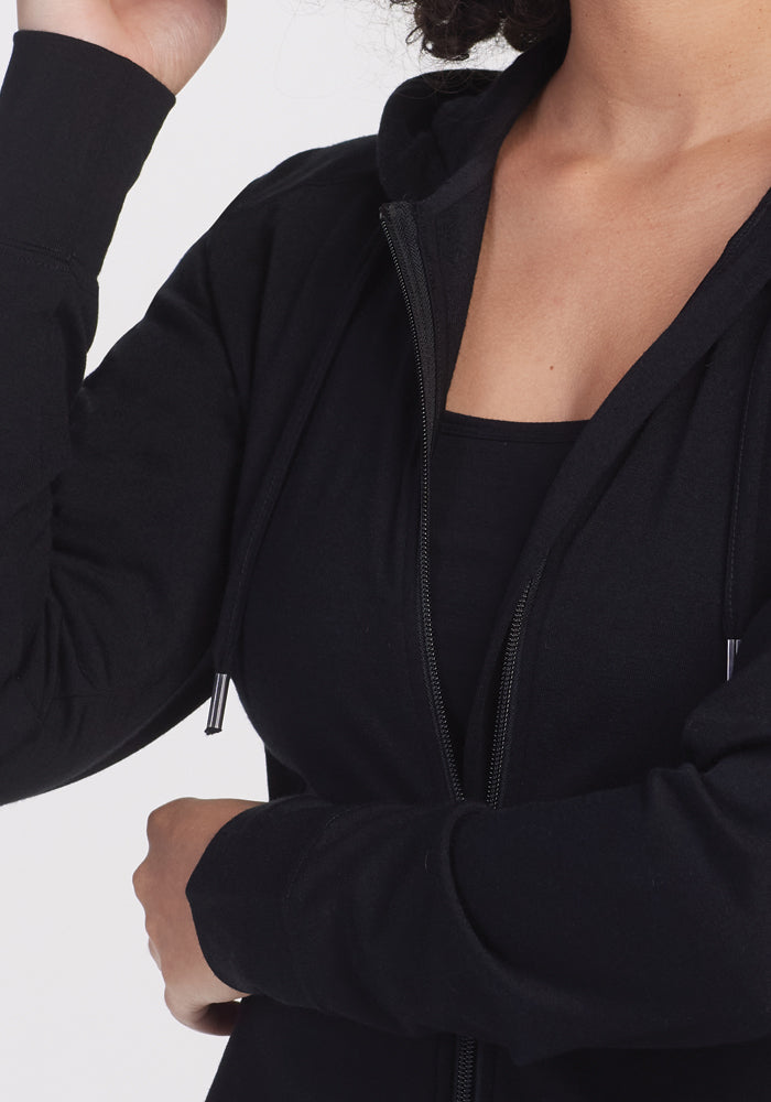 The Ryann Hoodie by Woolx, a lightweight black essential for travel, is worn from neck to waist with one arm slightly raised, set against a plain white background.