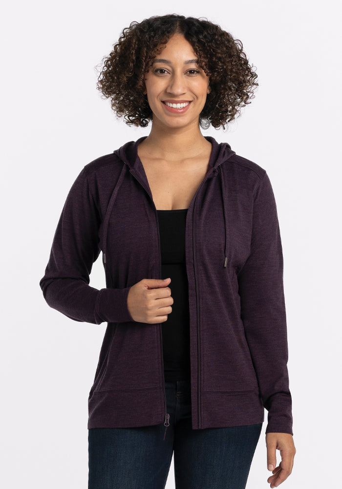 A person with curly hair is smiling, wearing the Woolx Ryann Hoodie in Deep Plum, which has a lightweight and athletic fit, over a black top and jeans. The background is plain white. 