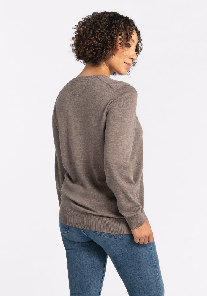 A person with curly hair is wearing a Francesca Sweater in Sable by Woolx and blue jeans, standing sideways and smiling against a plain white background.