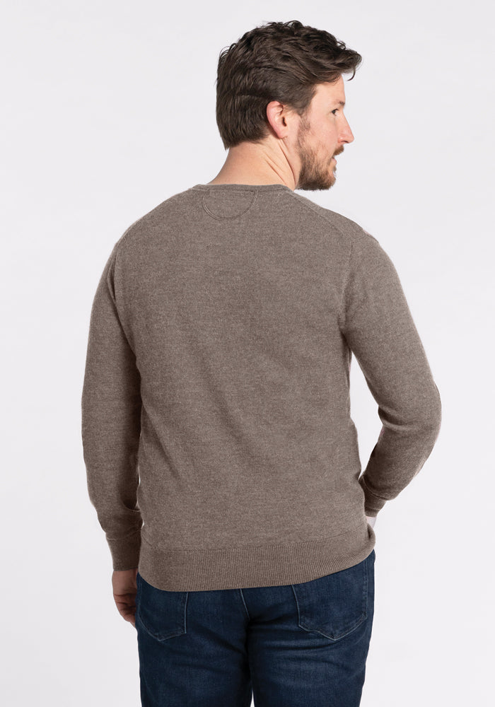 A man with short dark hair and a beard is wearing the Vincenzo Sweater in Sable by Woolx, along with dark jeans. He is standing and facing away from the camera against a plain white background.