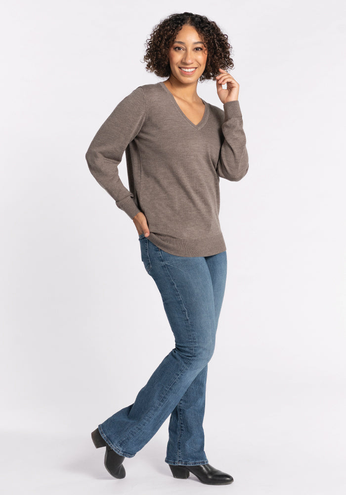 A person with curly hair stands smiling, wearing a Francesca Sweater in Sable by Woolx, along with blue jeans and black shoes. The person is posing with one hand in a pocket and the other touching their hair against a plain white background.