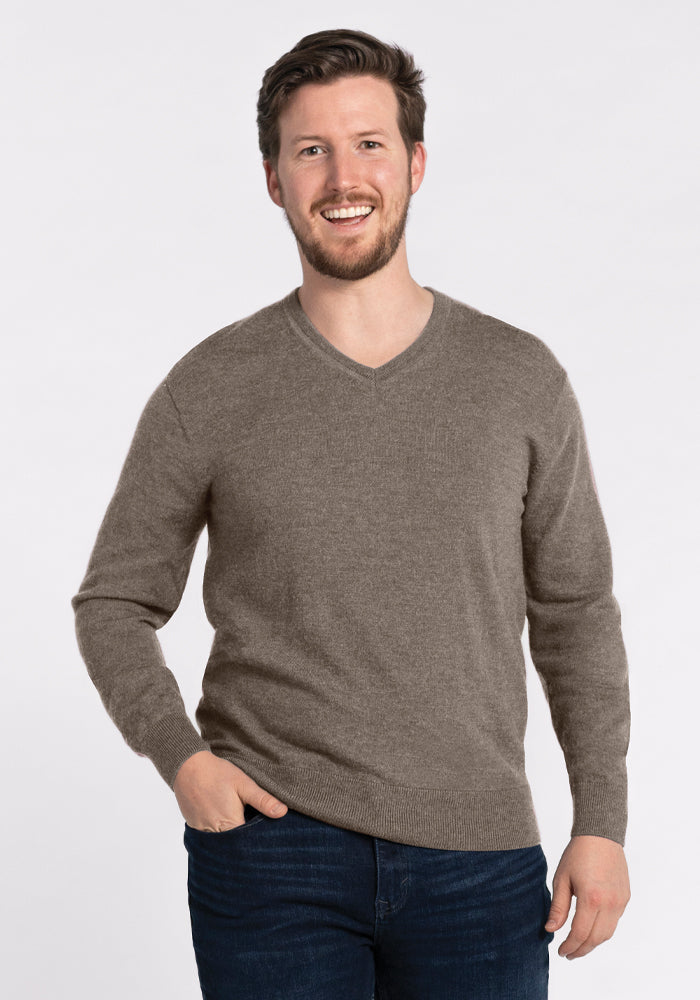 A man with short, brown hair and a beard is smiling while wearing the Sable Vincenzo Sweater by Woolx and dark jeans. He stands against a plain white background with one hand in his pocket. 