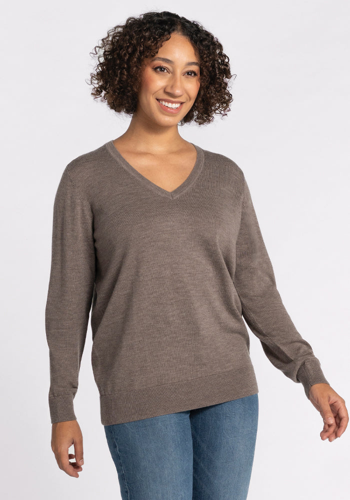 A person with curly hair smiles while wearing the Francesca Sweater in Sable by Woolx and blue jeans. The background is plain white.