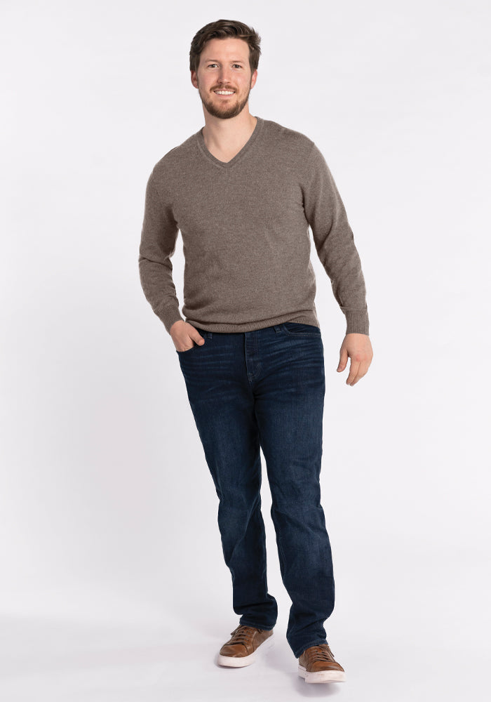 A bearded man is smiling and posing against a plain white background. He's wearing a Vincenzo Sweater in Sable by Woolx, dark blue jeans, and brown shoes. His left hand is in his pocket, and he seems to be stepping forward.