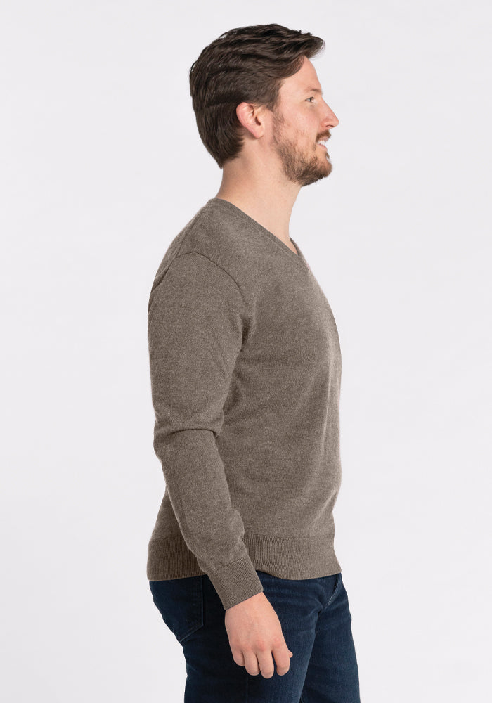 A man wearing the Vincenzo Sweater in Sable by Woolx and dark jeans stands in profile against a plain white background. His hands rest at his sides, and he looks forward.