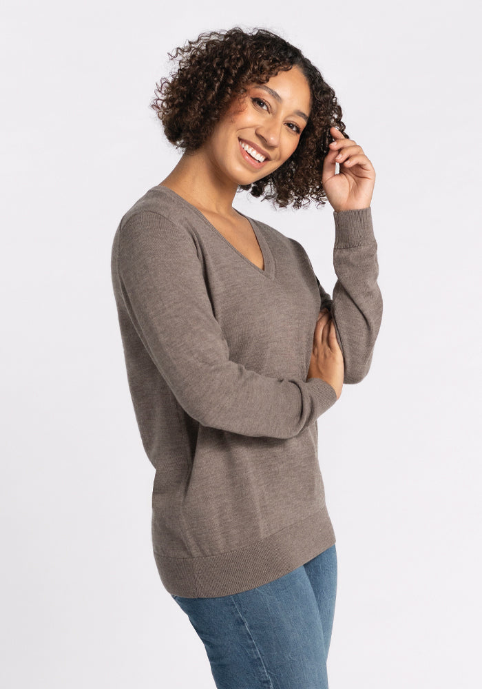 A person with curly hair smiles, wearing the Francesca Sweater in Sable from Woolx and blue jeans, standing against a plain white background. 