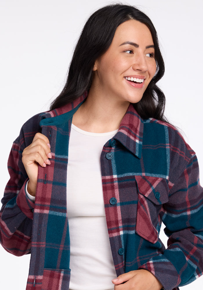 A smiling woman with long dark hair is wearing a Woolx Sawyer Shacket in blue, red, and white tones over a white T-shirt. The shacket features front pockets and large buttons. She is looking to her left and holding one side of her stylish jacket with her right hand.