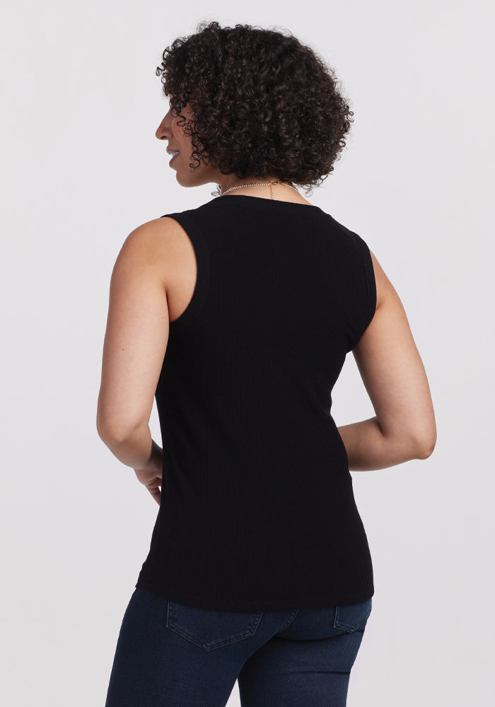 A person with curly hair, wearing a Woolx Sloane Ribbed Tank in black and dark jeans, stands with their back to the camera against a plain background.