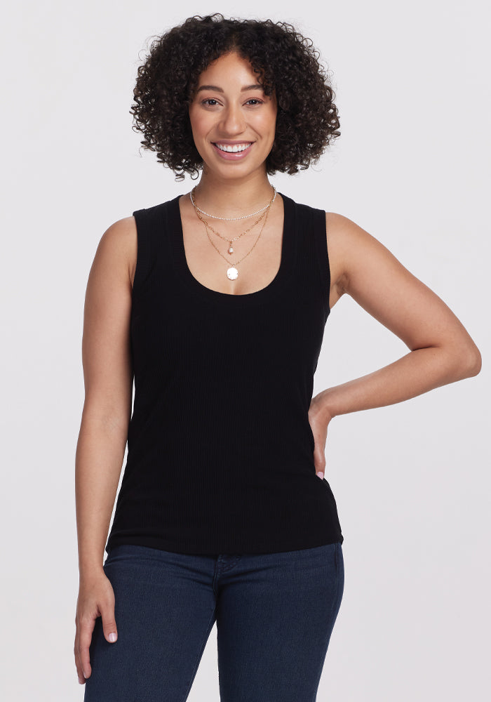 A person with curly hair smiles against a plain background, highlighting the versatile fit of their Sloane Ribbed Tank in black by Woolx. Paired with blue jeans and layered necklaces, it creates a stylish yet effortless look. 