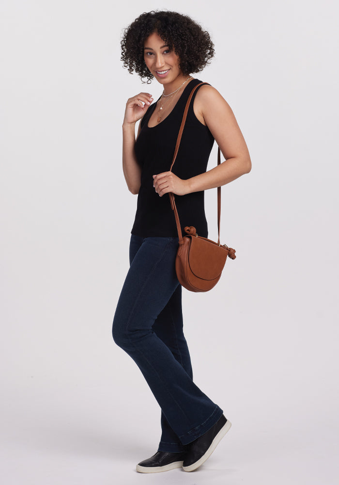 A curly-haired person smiles on a plain white background, wearing a versatile Black Sloane Ribbed Tank by Woolx paired with blue jeans. They hold a brown crossbody bag and sport black slip-on shoes crafted from luxurious Australian Merino Wool.