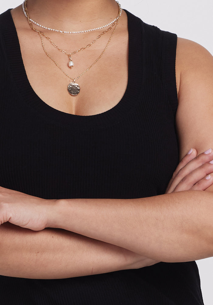 A person in a Sloane Ribbed Tank - Black by Woolx has arms crossed, showcasing layered gold necklaces with a small pendant and pearl. The image emphasizes the fit of the top and jewelry, focusing on the upper torso while keeping the face hidden.