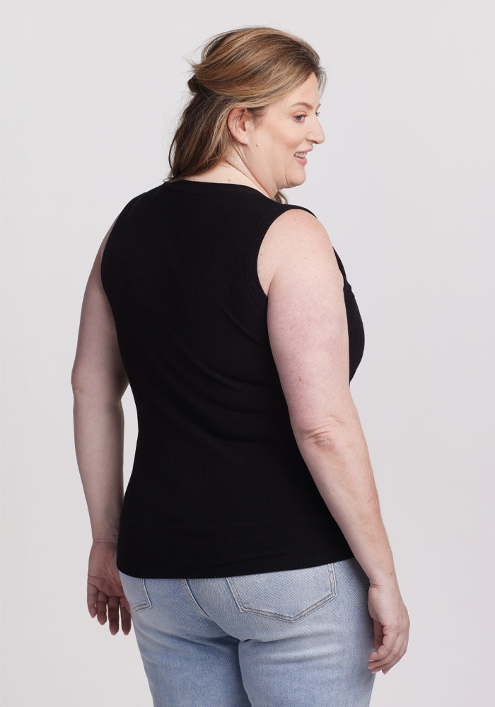 Against a plain gray background, a person with long hair smiles in a versatile Woolx Sloane Ribbed Tank - Black and light blue jeans, turned slightly to the side.