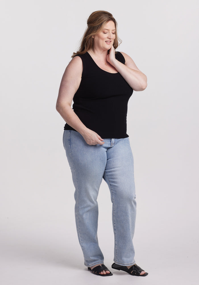 A woman wearing a black Woolx Sloane Ribbed Tank, light blue jeans, and black sandals stands against a plain white background. With one hand on her neck and the other on her hip, she smiles slightly, highlighting the versatile fit of her outfit.