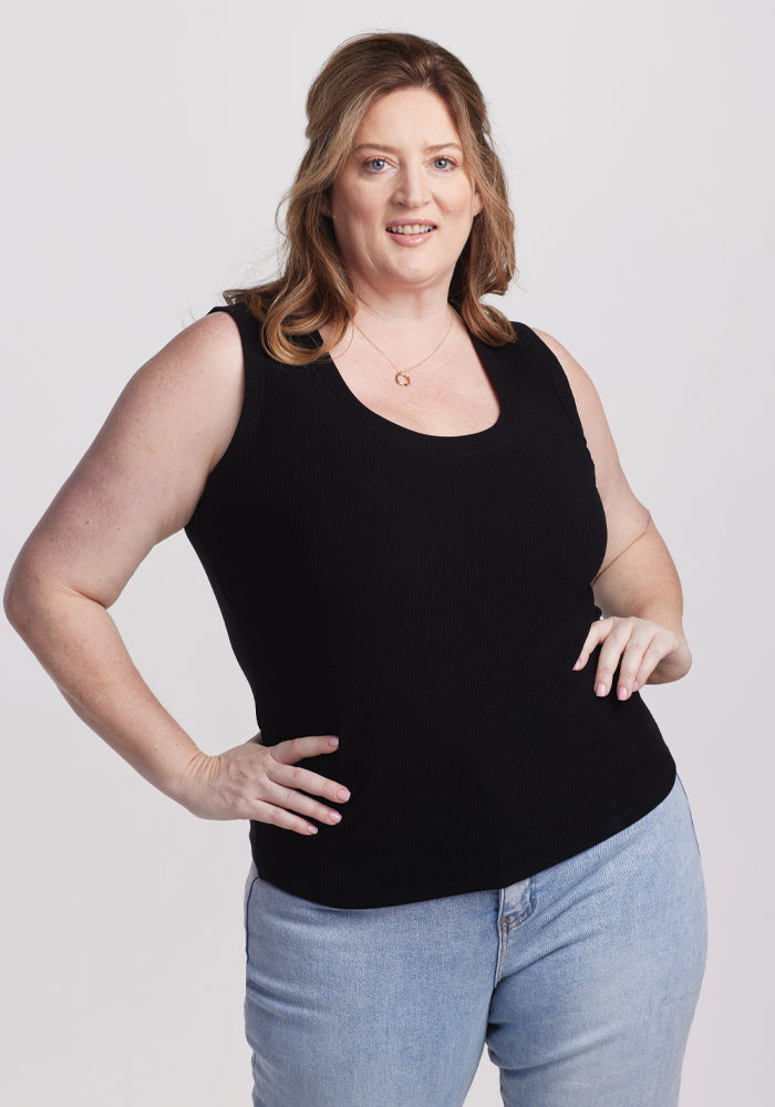 A person with long hair smiles with one hand on their hip against a plain white background, wearing a Woolx Sloane Ribbed Tank in black and light blue jeans. 