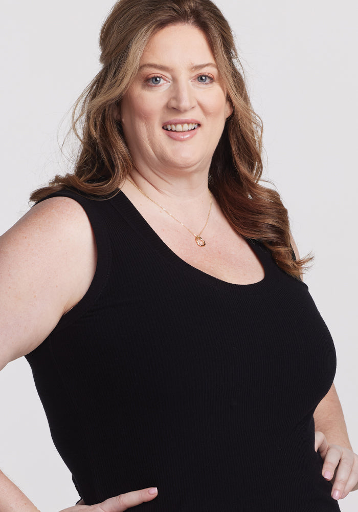 A person with long brown hair smiles, wearing a Woolx Sloane Ribbed Tank in black, highlighting its versatile fit and paired with a gold necklace against a plain light-colored background.