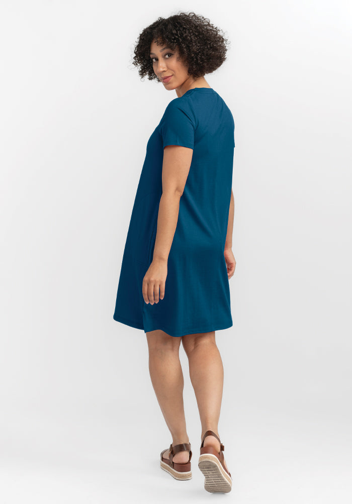 A person with curly hair is wearing the Georgie Dress in Real Teal by Woolx, a short-sleeved A-line dress crafted from lightweight fabric, and paired with brown sandals. They are positioned on a white background, turned slightly and glancing over their shoulder.