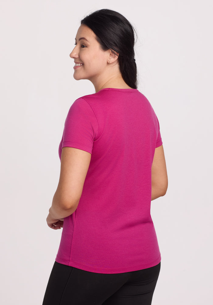 A smiling person with long dark hair is seen in profile, slightly turned back, wearing a bright pink Liza Crew Neck Tee - Berry Punch by Woolx and black pants against a plain white background.