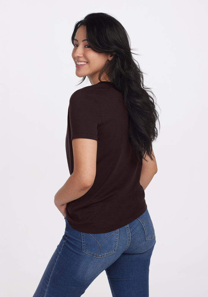 A person with long dark hair stands smiling, wearing the Woolx Liza Crew Neck Tee in French Roast, crafted from lightweight fabric, paired with blue jeans. They are looking over their shoulder against a plain white background.