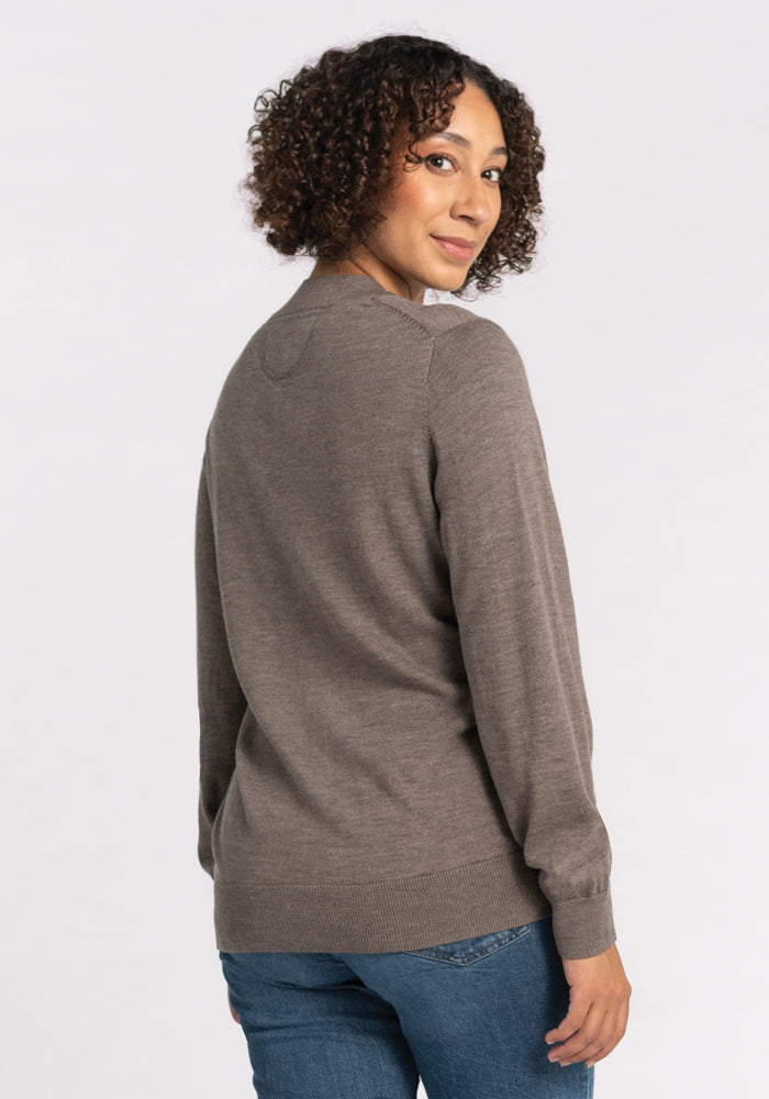 A person with curly hair is slightly turning towards the camera, dressed in a brown Gia Sweater - Sable from Woolx and blue jeans, standing against a plain background. This travel essential promises temperature-regulating comfort wherever your journey takes you.