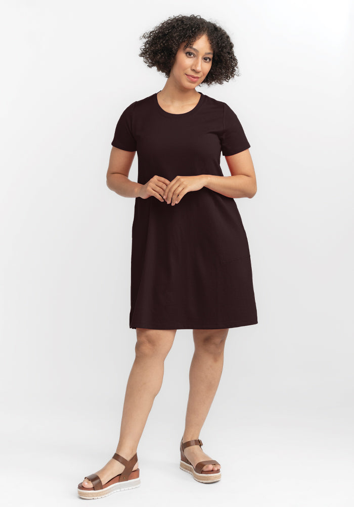 A person with curly hair stands against a white background, wearing a short-sleeved French Roast Georgie Dress by Woolx and brown wedge sandals. They have a relaxed posture, with one hand lightly touching the other. 