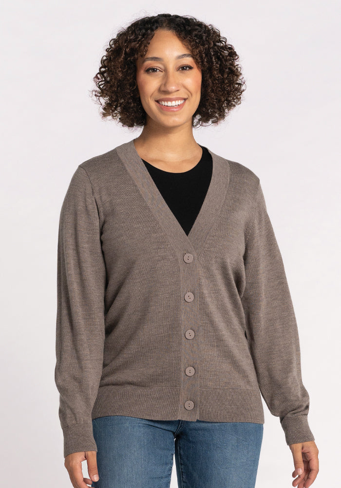 A person with curly hair smiles while wearing the Gia Sweater in Sable from Woolx over a black shirt and blue jeans, standing against a neutral background. This travel essential offers temperature-regulating comfort, making it perfect for any journey. 