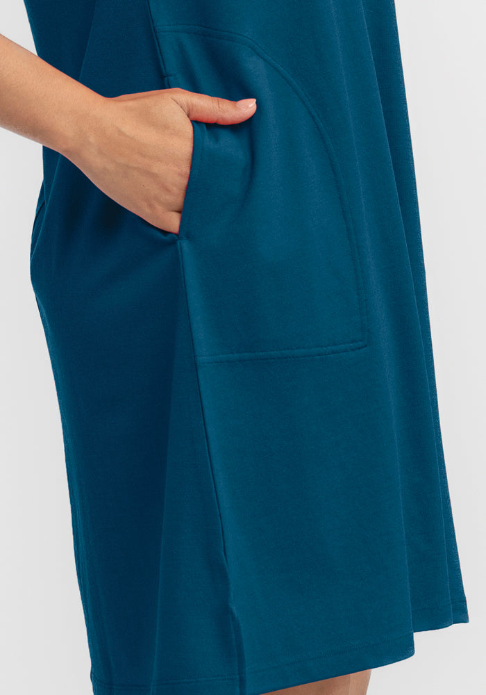 A person clad in the Woolx Georgie Dress - Real Teal, featuring an A-line silhouette with a noticeable side pocket, slips their left hand inside. Made from a lightweight fabric, the dress elegantly drapes on the body against a plain white backdrop.