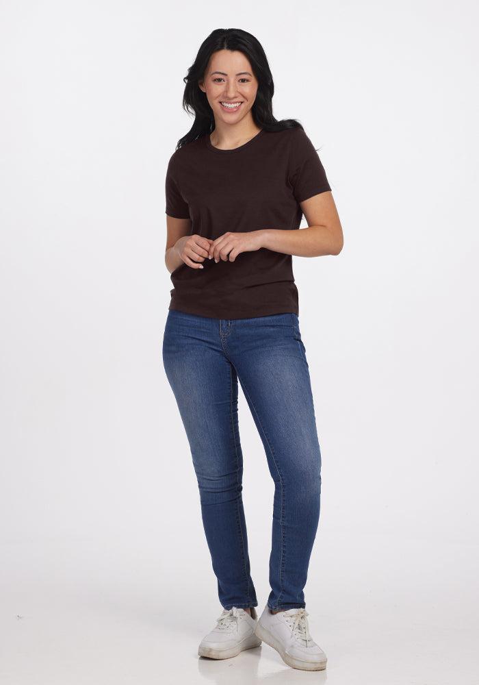 A person with long dark hair stands smiling against a white background, wearing the Liza Crew Neck Tee in French Roast by Woolx, along with blue jeans and white sneakers. Their hands are relaxed in front of them.