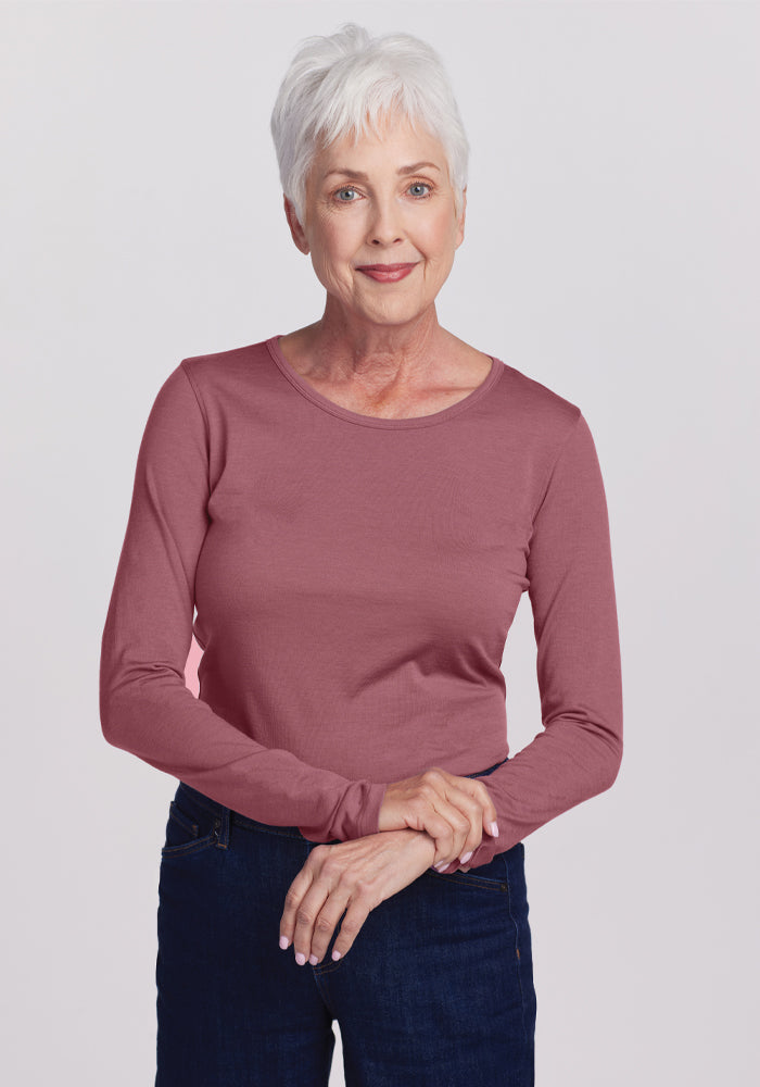 An older woman with short white hair is wearing the Woolx Remi Long Sleeve T-Shirt in Wild Ginger along with dark jeans, standing against a plain white background. She smiles gently and holds her hands together in front of her. 