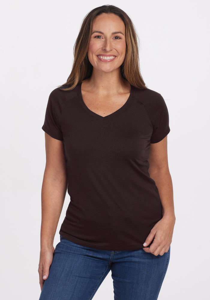 A smiling woman with long hair stands against a plain background, wearing the Mia V Neck in French Roast from Woolx and blue jeans. Her right hand rests on her hip while her left arm hangs by her side, exuding an effortlessly chic, odor-free vibe. 