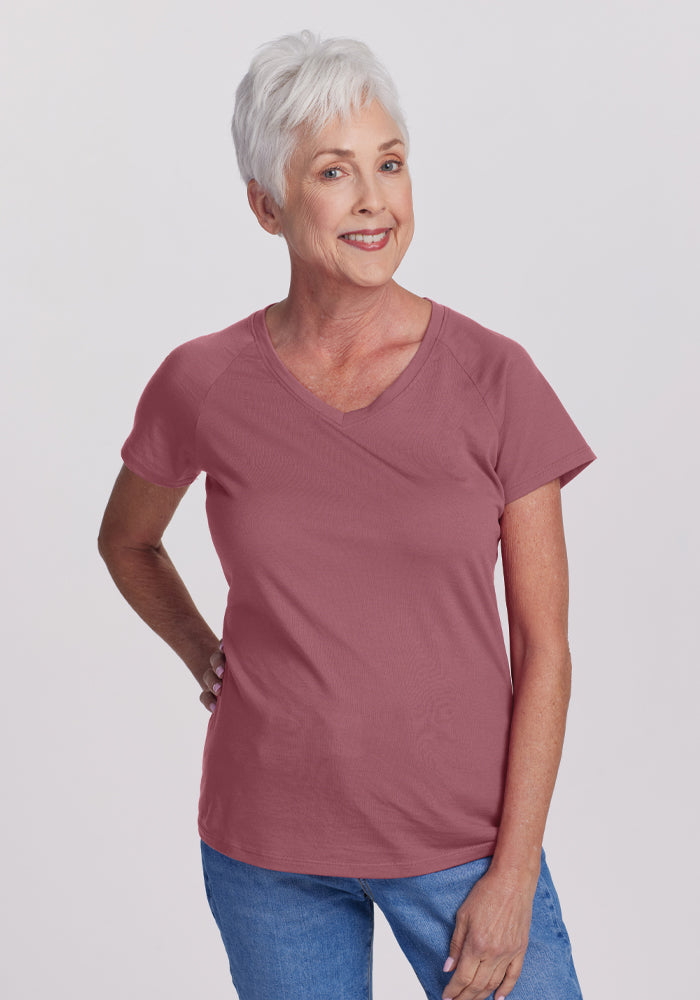 An older woman with short white hair smiles warmly, dressed in a Wild Ginger Mia V Neck by Woolx. She pairs it with blue jeans as she stands against a plain white background, her right hand confidently on her hip. 