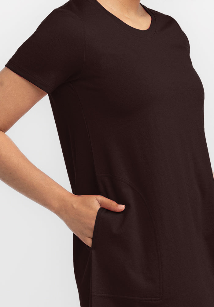 A person wearing a French Roast Georgie Dress from Woolx, crafted from merino wool, stands against a white background. The A-line dress includes a convenient pocket where the person's left hand rests comfortably. Only the torso and upper arm are visible.