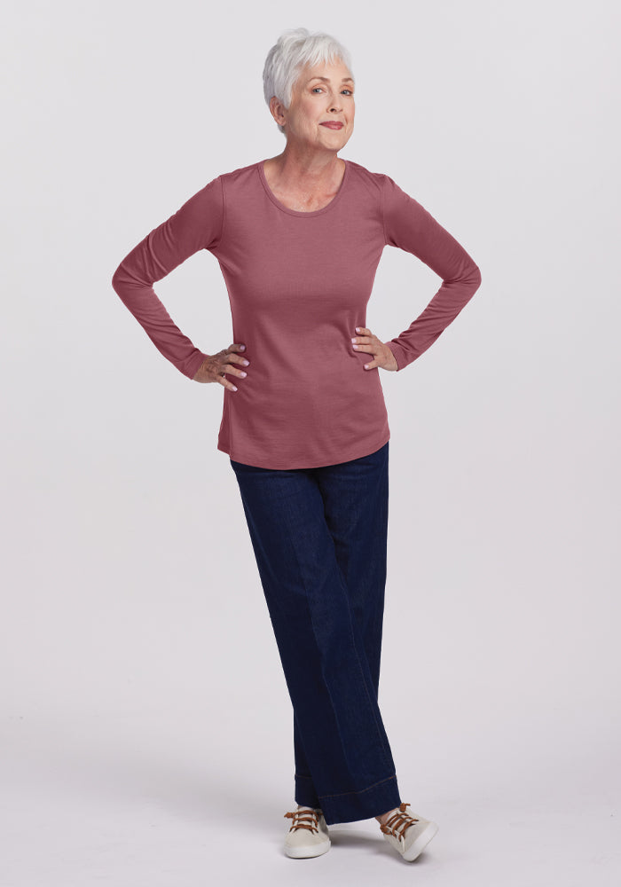 An older adult with short white hair poses in a relaxed manner, highlighting the comfort of their Remi Long Sleeve T-Shirt in the Wild Ginger color from Woolx. Paired with blue jeans and white shoes, they stand against a plain white background.