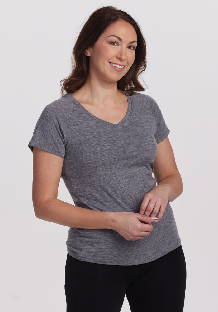 Wearing a comfortable Woolx Mia V Neck in Graphite Heather and black pants, a person smiles while standing against a plain light background, casually holding their hands together in front. 