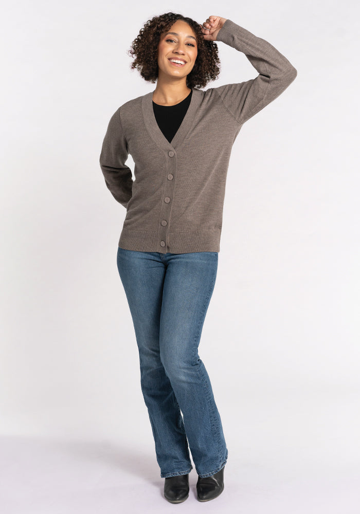 A person poses confidently with one hand on their head and the other in their pocket, showcasing the Gia Sweater - Sable by Woolx. This travel essential offers temperature-regulating comfort and is styled with a black top, blue jeans, and black shoes against a plain white background.