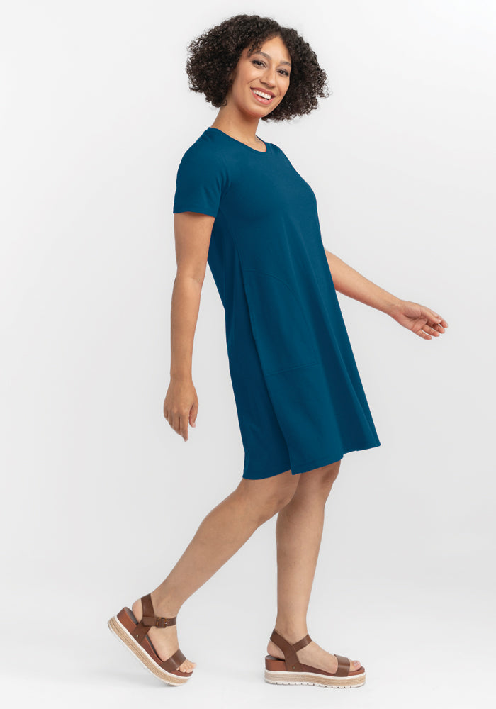 A person with curly hair is smiling and walking sideways, dressed in a relaxed-fit Georgie Dress in Real Teal from Woolx, made of soft merino wool. They pair it effortlessly with brown platform sandals against a plain white studio backdrop.