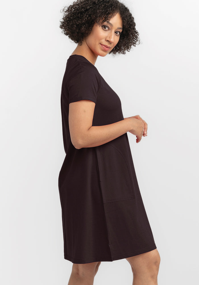 A person with curly hair is elegantly wearing a French Roast-colored Georgie Dress by Woolx, standing sideways and looking at the camera. The background is a crisp white.