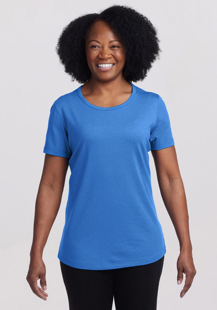 A curly-haired person smiles in a bright blue Liza Crew Neck Tee - Blue Gelato from Woolx, paired with black pants, against a light gray background. 