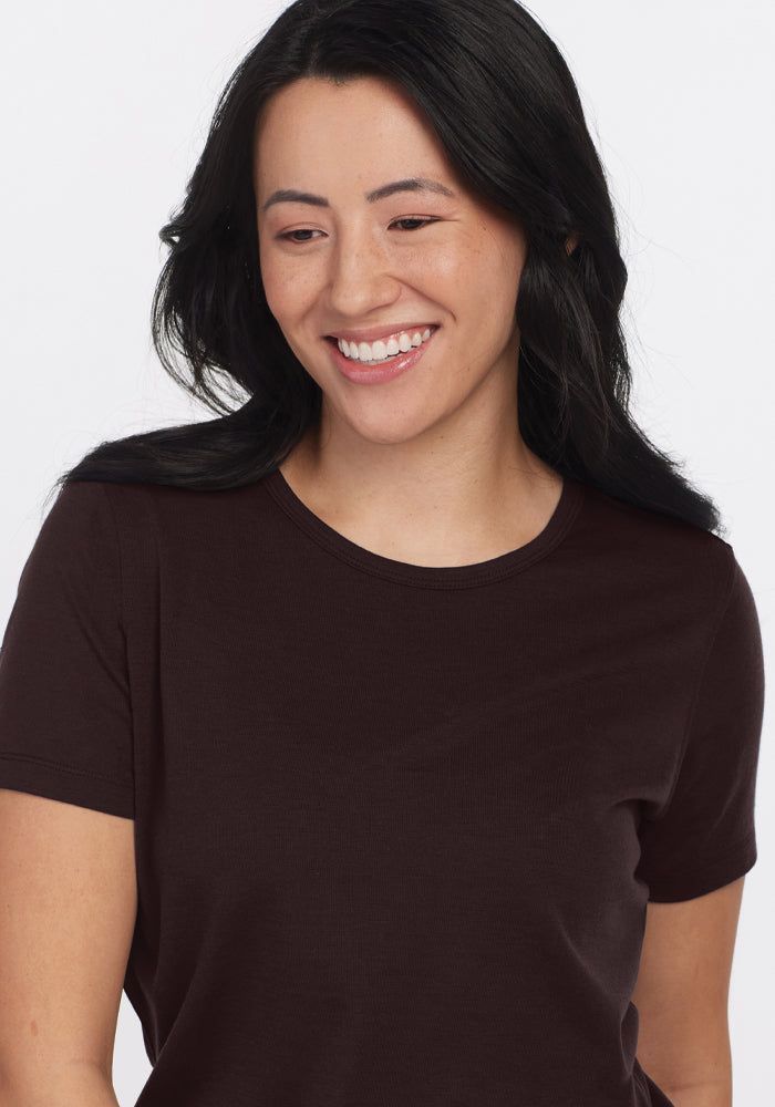 A person with long black hair is smiling and looking slightly to the side, wearing a Woolx Liza Crew Neck Tee in French Roast, made from lightweight fabric, against a plain white background.