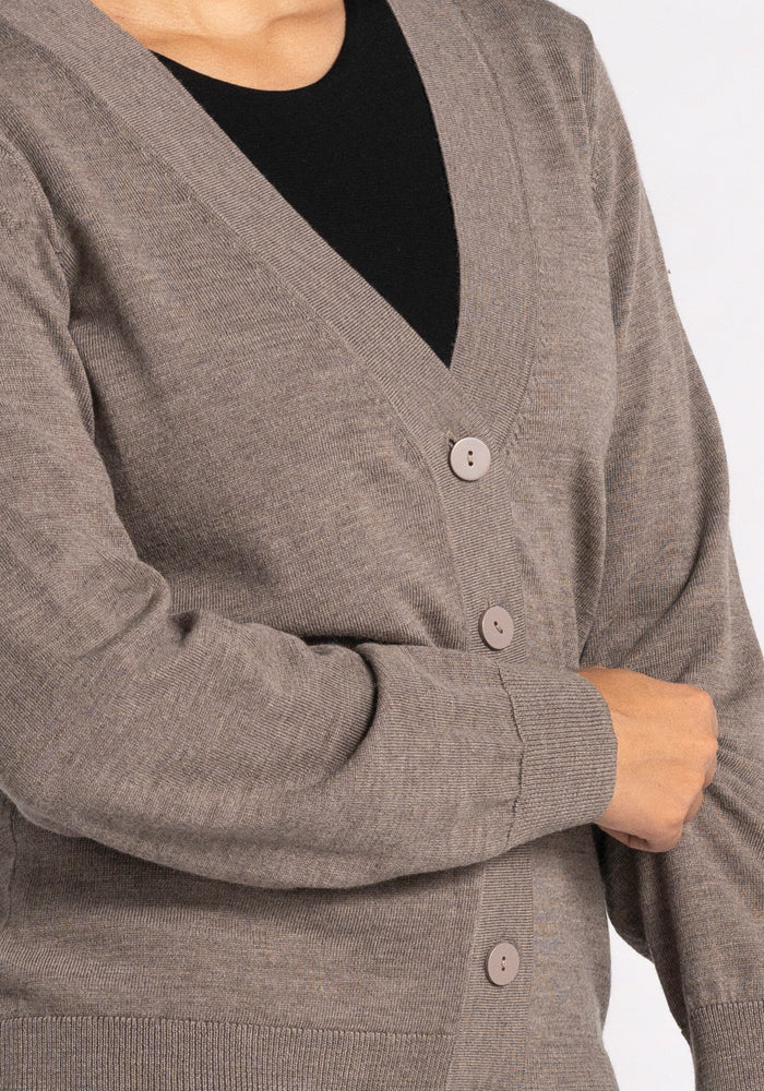 A person wearing the Gia Sweater - Sable over a black shirt. Crafted from Merino wool by Woolx, this lightweight cardigan features a V-neck and is fastened with three round buttons. The person's hand rests confidently on their hip, embodying the perfect travel companion look.