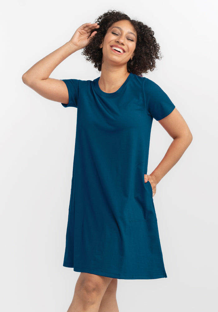 A person with curly hair beams joyfully while wearing the Georgie Dress - Real Teal by Woolx, a short-sleeved A-line dress with pockets. Made from a lightweight fabric, they pose against a plain white background, one hand raised to their head.