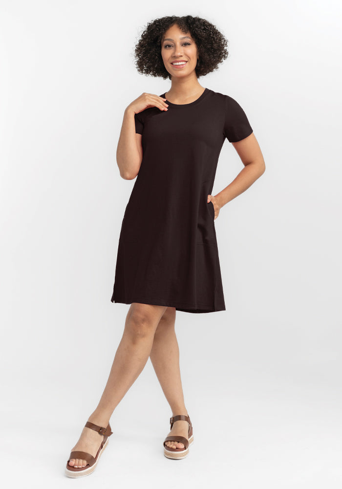 A person with curly hair smiles while posing in a French Roast Georgie Dress by Woolx and tan sandals. They are standing against a plain white background, one hand on their hip and the other touching their chin.