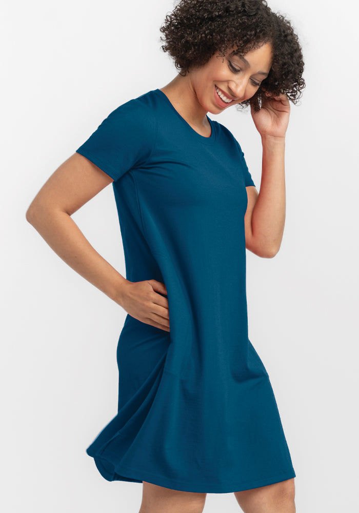 A person with curly hair is smiling and wearing the Georgie Dress in Real Teal by Woolx. They are posing with one hand on their hip and the other touching their hair against a plain white background.