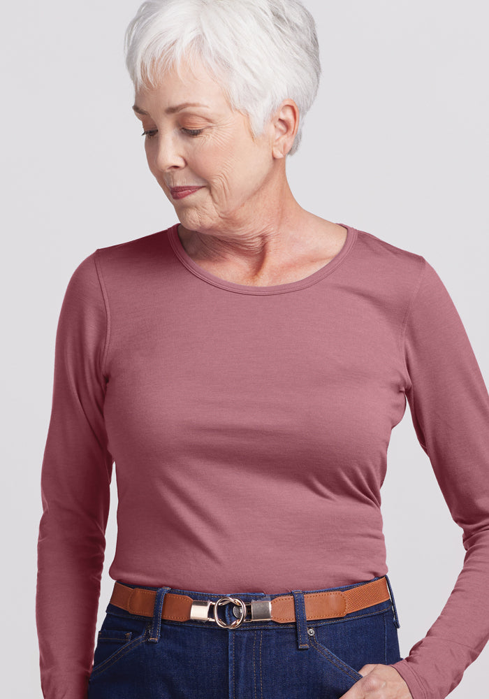 An older woman with short white hair is wearing a Remi Long Sleeve T-Shirt in Wild Ginger by Woolx, made from ultra-lightweight fabric, along with dark jeans and a brown belt. She gazes downward with a neutral expression against a plain background.