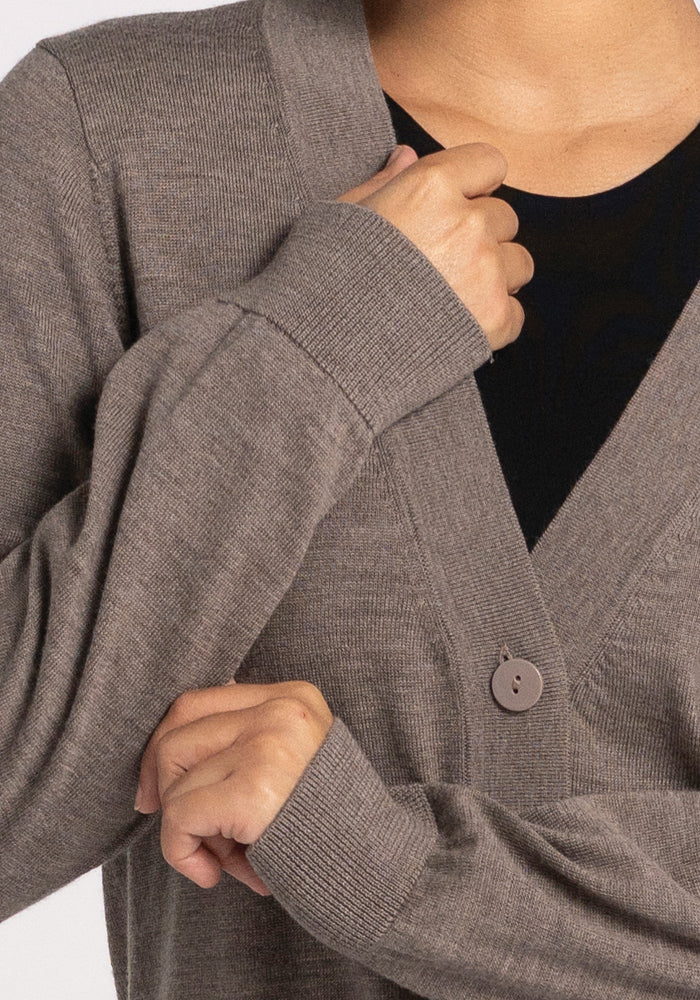 A person is wearing the Gia Sweater - Sable by Woolx over a black top, with one hand grasping the collar and the other resting on their arm. This travel essential sweater features a visible button and provides temperature-regulating comfort against a plain white background.