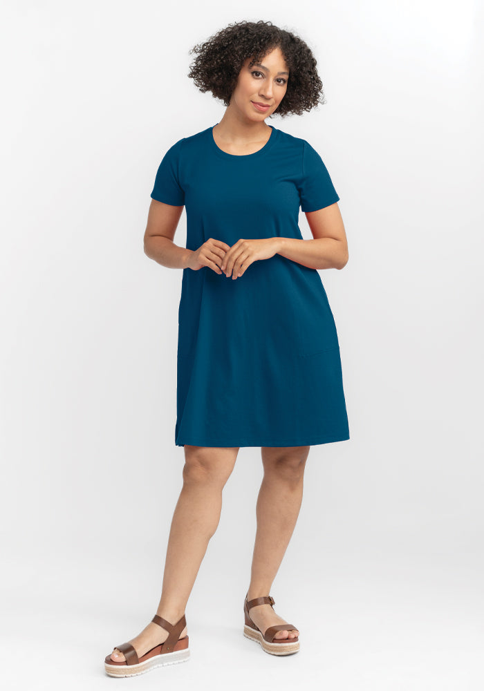 A person with curly hair stands against a plain white background, wearing the Woolx Georgie Dress in Real Teal, crafted from lightweight fabric. They complement the outfit with brown platform sandals while gazing at the camera with a slight smile and a relaxed posture.