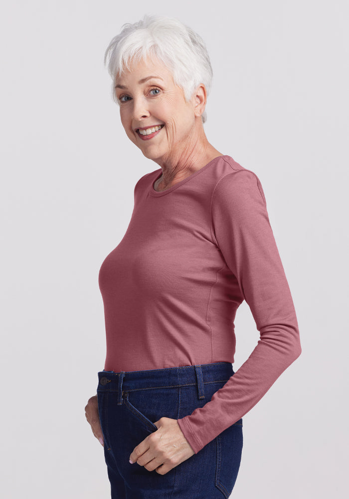 An older woman with short white hair smiles while standing sideways, wearing the Remi Long Sleeve T-Shirt in Wild Ginger by Woolx, paired with dark blue jeans. The lightweight fabric of her shirt complements her relaxed demeanor against the plain light background.