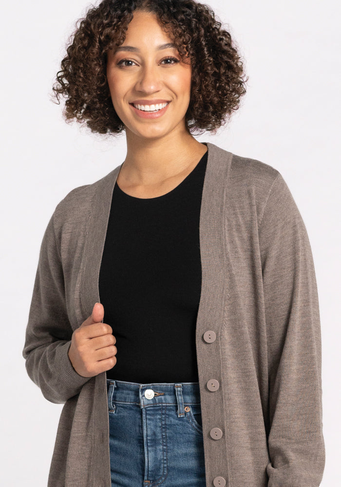 A person with curly hair smiles, enjoying the temperature-regulating comfort of their Gia Sweater in Sable, crafted from Merino wool by Woolx. Paired with a black shirt and blue jeans, they stand against a plain, light background—a true travel essential for any journey.