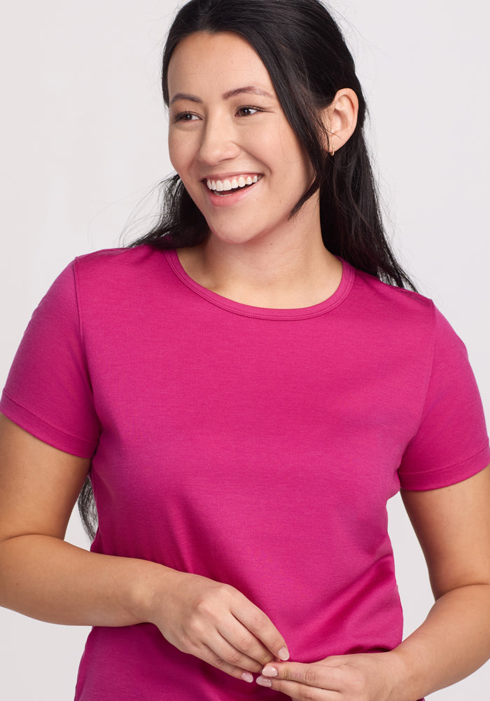 A person with long dark hair smiles and looks to the side while wearing a Liza Crew Neck Tee - Berry Punch by Woolx, crafted from lightweight fabric. The background is plain and light-colored.